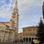 Tall bell tower beside historic buildings and Christmas tree in Modena, Italy under a blue sky.