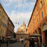 Historic street view with people, colorful buildings, and a grand church in Modena, Italy, under a clear sky.