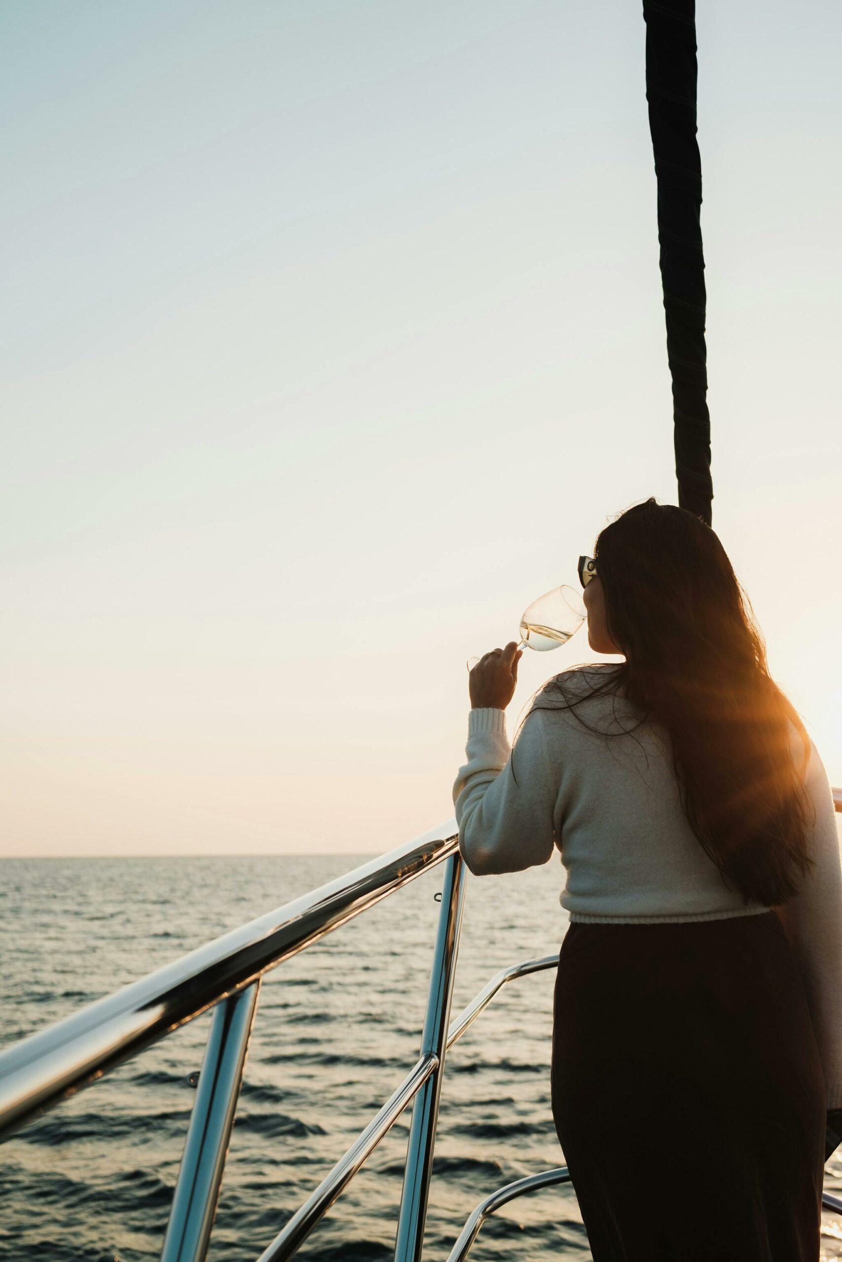Woman enjoying a sunset view on a yacht, sipping wine, with ocean horizon and warm sunlight.